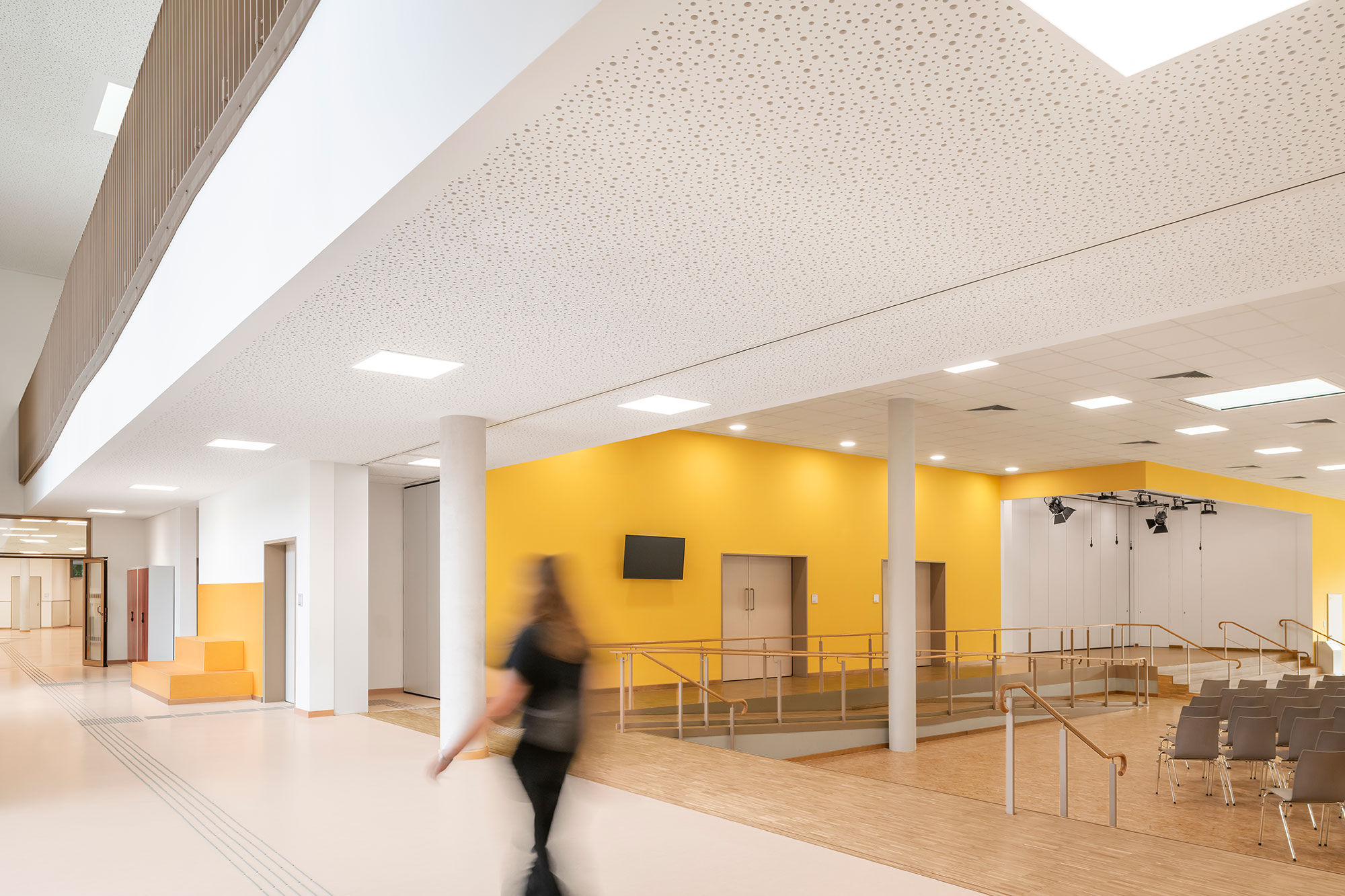 Aula und Mensa der neu errichteten Grundschule Mühlenberg mit heller, freundlicher Gestaltung: ein großzügiger Eingangsbereich mit gelber Akzentwand, barrierefreier Rampe, Sitzstufen und Decke mit runden Akustiköffnungen; rechts befindet sich ein  Veranstaltungsbereich mit Bühne und Stuhlreihen, im Vordergrund geht eine unscharf dargestellte Person vorbei.