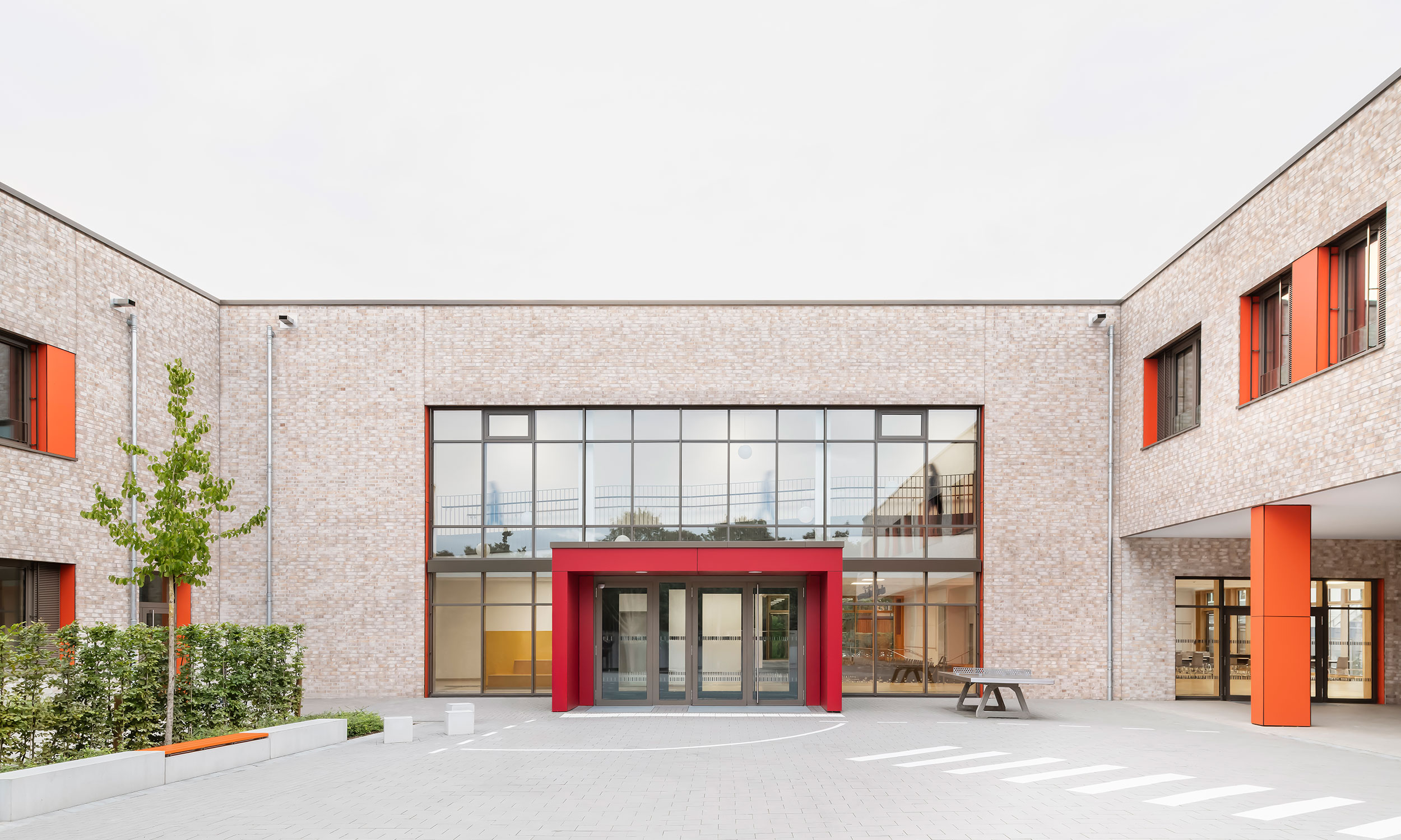 Rückansicht der Grundschule Mühlenberg vom Schulhof aus, mit einer zweigeschossigen hellen Klinkerfassade, großen Fensterfronten und einem markanten roten Eingangsbereich.