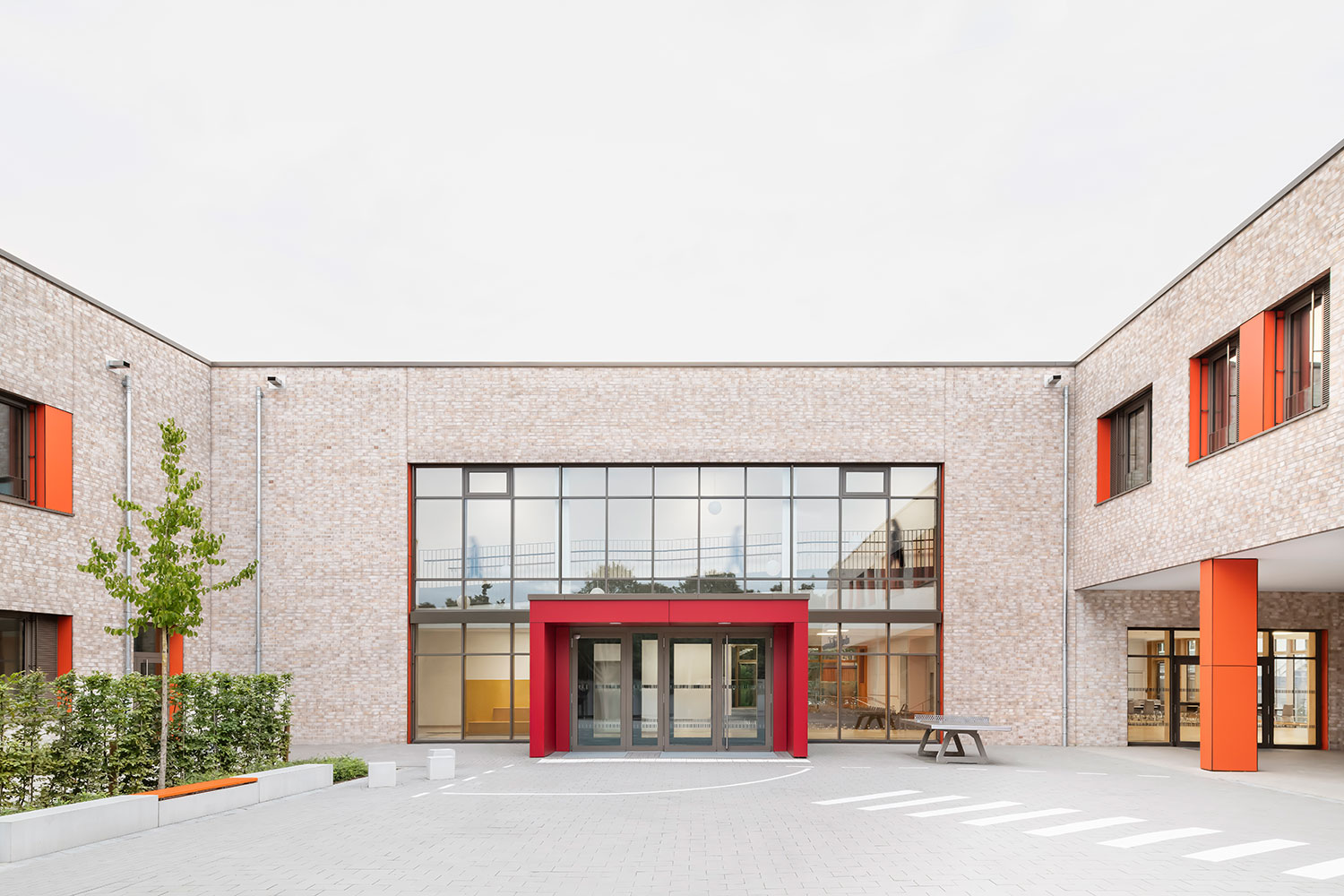 Rückansicht der Grundschule Mühlenberg vom Schulhof aus, mit einer zweigeschossigen hellen Klinkerfassade, großen Fensterfronten und einem markanten roten Eingangsbereich.