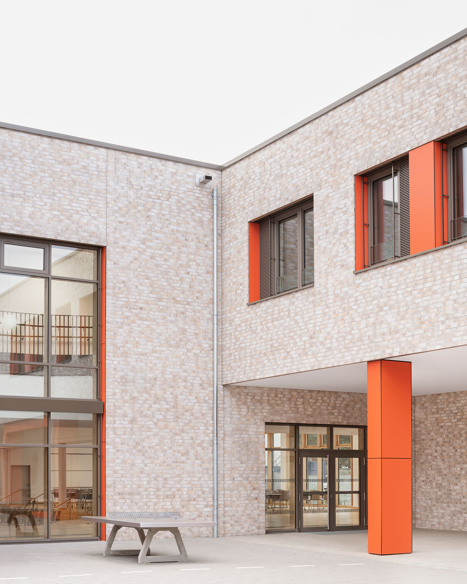 Detailansicht der neu errichteten Grundschule Mühlenberg vom Schulhof aus, mit heller Klinkerfassade, großformatigen Fenstern und markanten orangefarbenen Fassadenpaneelen und Stützen; im Vordergrund steht eine Tischtennisplatte, während im Hintergrund durch die Glasfront ein Innenraum sichtbar ist.