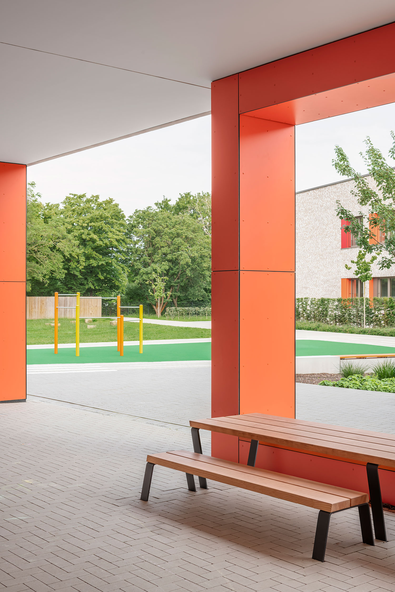 Detailansicht eines überdachten Bereichs der Grundschule Mühlenberg, mit orangefarben verkleideten Stützen und einer Holzsitzbank im Vordergrund; durch die Öffnung sind der grüne Außenbereich mit Spielgeräten sowie die helle Klinkerfassade des Schulgebäudes sichtbar.