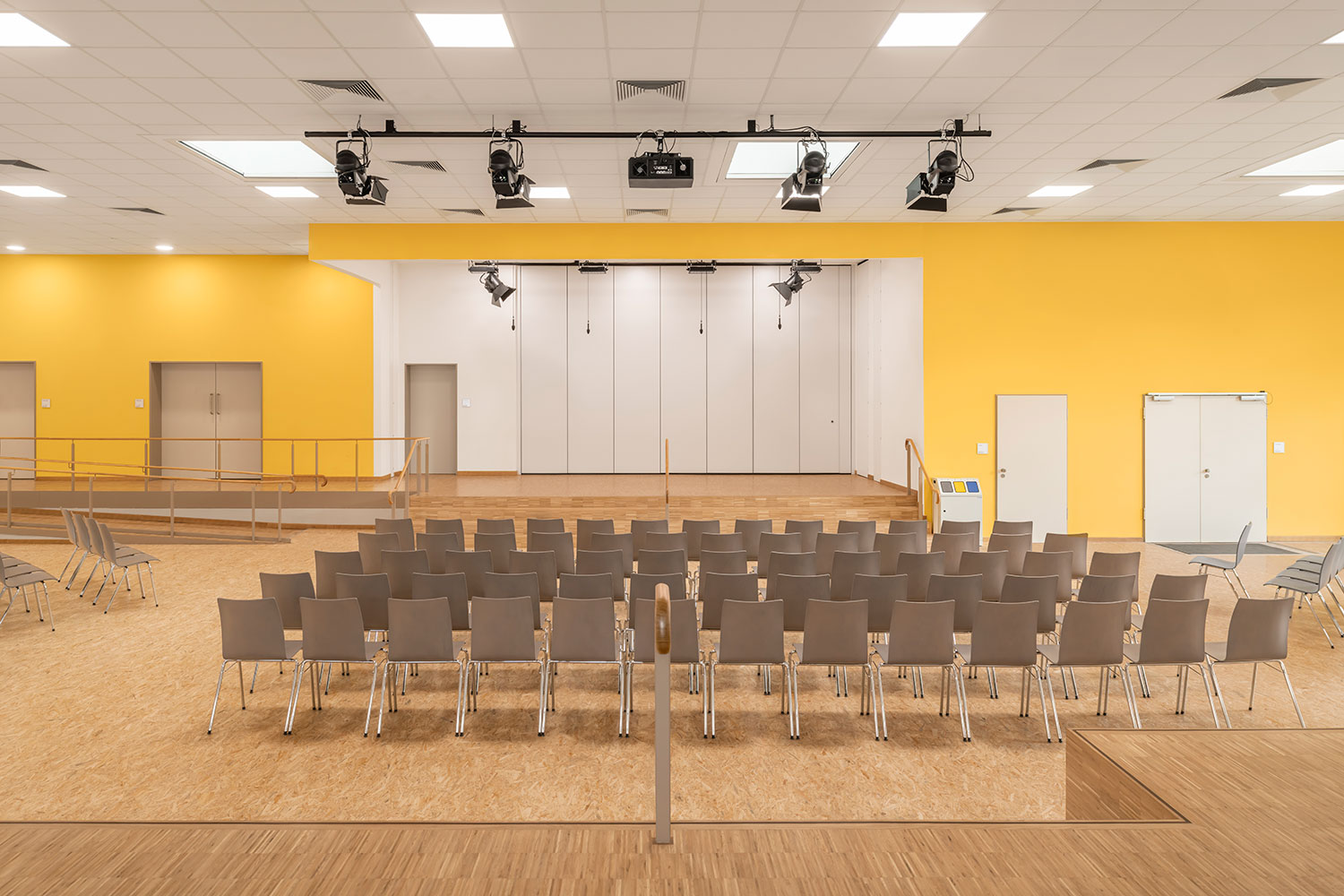 Aula der neu errichteten Grundschule Mühlenberg mit heller gelber Akzentwand, aufgestellten Stuhlreihen und einer erhöhten Bühne; über der Bühne sind Licht- und Technikspots installiert, dahinter befindet sich eine großflächige, flexible Trennwand zur variablen Raumteilung.