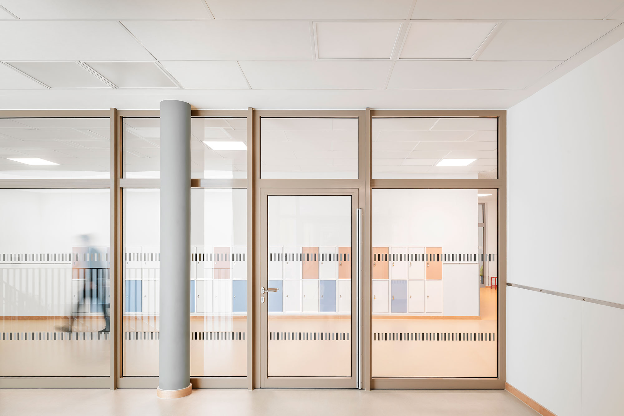 Verglaste Trennwand der neu errichteten Grundschule Mühlenberg mit einer zentralen Tür und großzügigen Glaselementen, durch die farbige Schließfächer sichtbar sind.