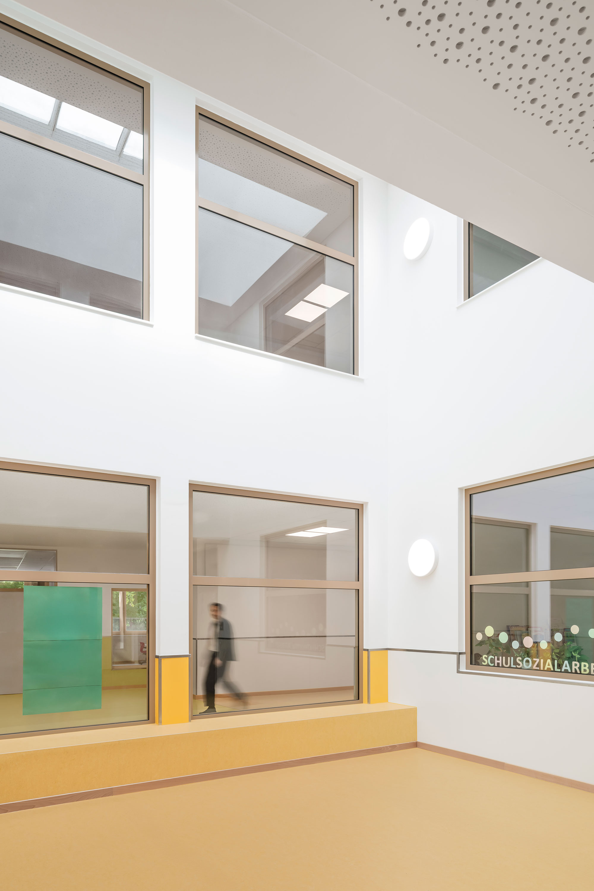 Innenraum der neu errichteten Grundschule Mühlenberg mit großformatigen Innenfenstern zu den Aufenthaltsbereichen im Ober- und Untergeschoss; die Wände sind weiß mit gelben Akzentbereichen, und durch die Scheiben ist eine unscharf vorbeigehende Person zu erkennen.