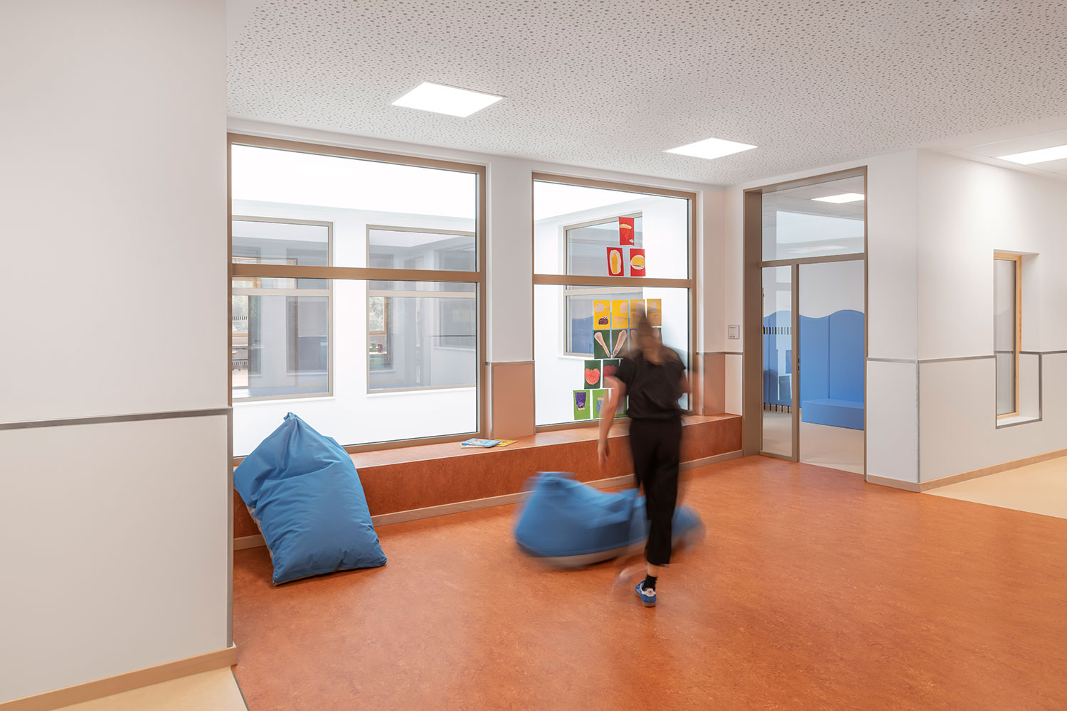 Aufenthalts- und Lernbereich der neu errichteten Grundschule Mühlenberg mit orangem Bodenbelag, breiter Fenstersitzbank und zwei blauen Sitzsäcken; durch die großen Innenfenster fällt viel Licht in den Raum, während eine unscharf dargestellte Person einen Sitzsack bewegt und farbige Kinderzeichnungen an der Glasscheibe hängen.