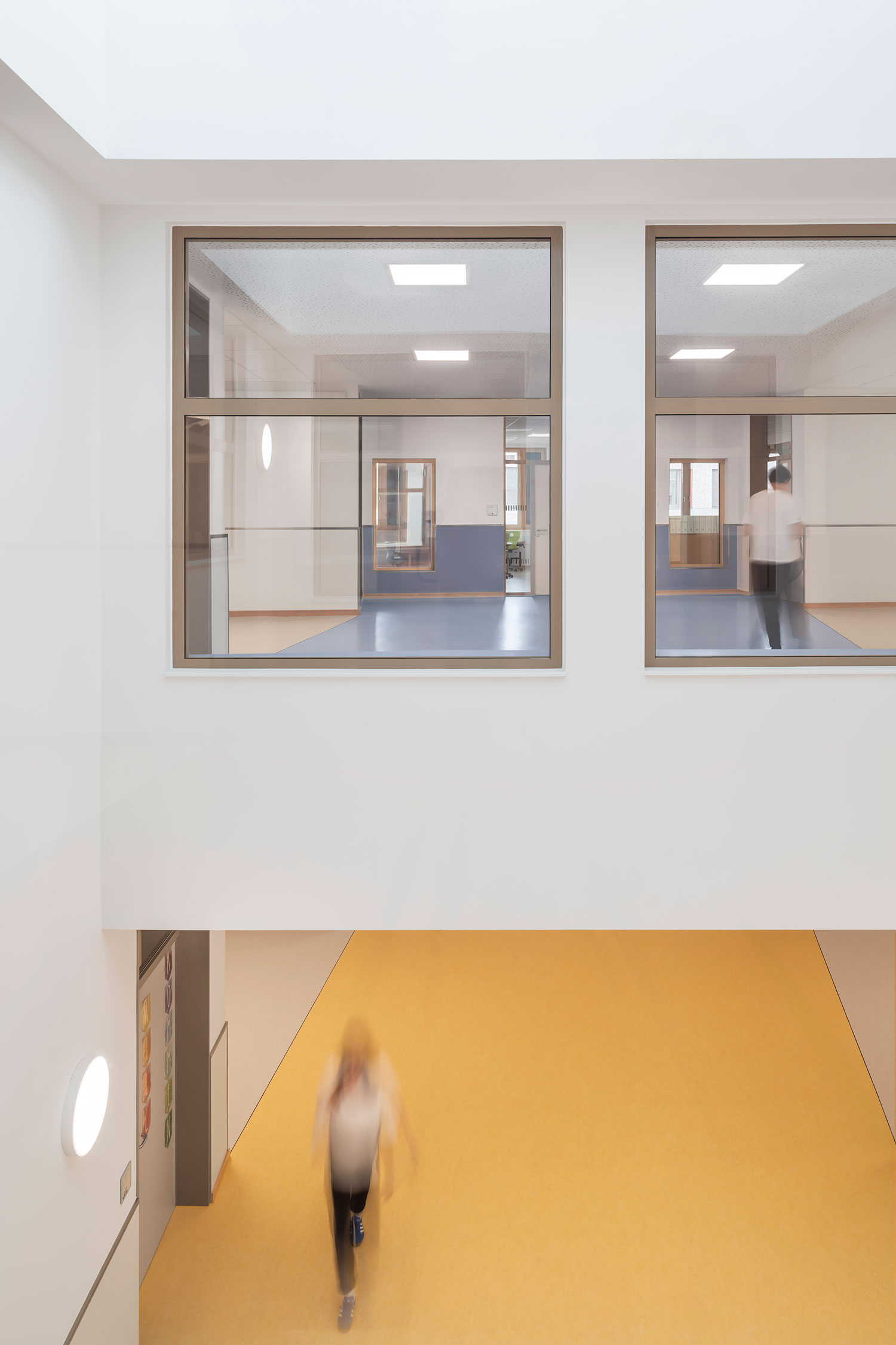 Blick in das zweigeschossige Atrium der neu errichteten Grundschule Mühlenberg, mit großen Innenfenstern zu den blau-weiß gestalteten Fluren im Obergeschoss; unten führt ein Bereich mit gelbem Bodenbelag vorbei, in dem eine unscharf vorbeilaufende Person zu sehen ist.