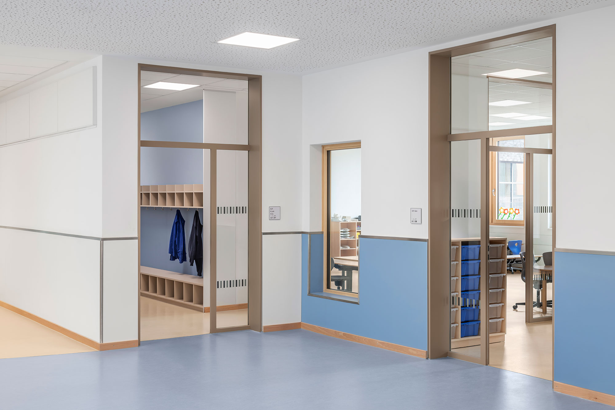 Lernwelt der neu errichteten Grundschule Mühlenberg mit hellblauer Wandgestaltung im unteren Bereich, großen Glaselementen, Sichtbeziehungen und offenen Zugängen zu Klassen- und Garderobenräumen.