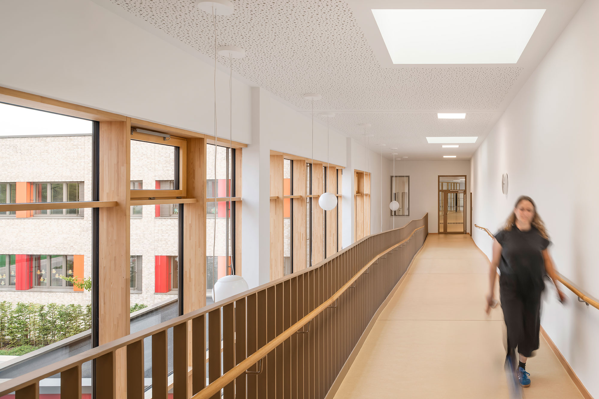 Lichtdurchflutetes Obergeschoss der neu errichteten Grundschule Mühlenberg mit einer offenen Galerie vor langen bodentiefen Holzrahmenfenstern, einem warmtonigen Metallgeländer und hellen Wand- und Deckenflächen mit Akustikperforation; links fällt der Blick auf die farbigen Fassadenelemente des Innenhofs, während rechts eine unscharf gehende Person zu sehen ist.