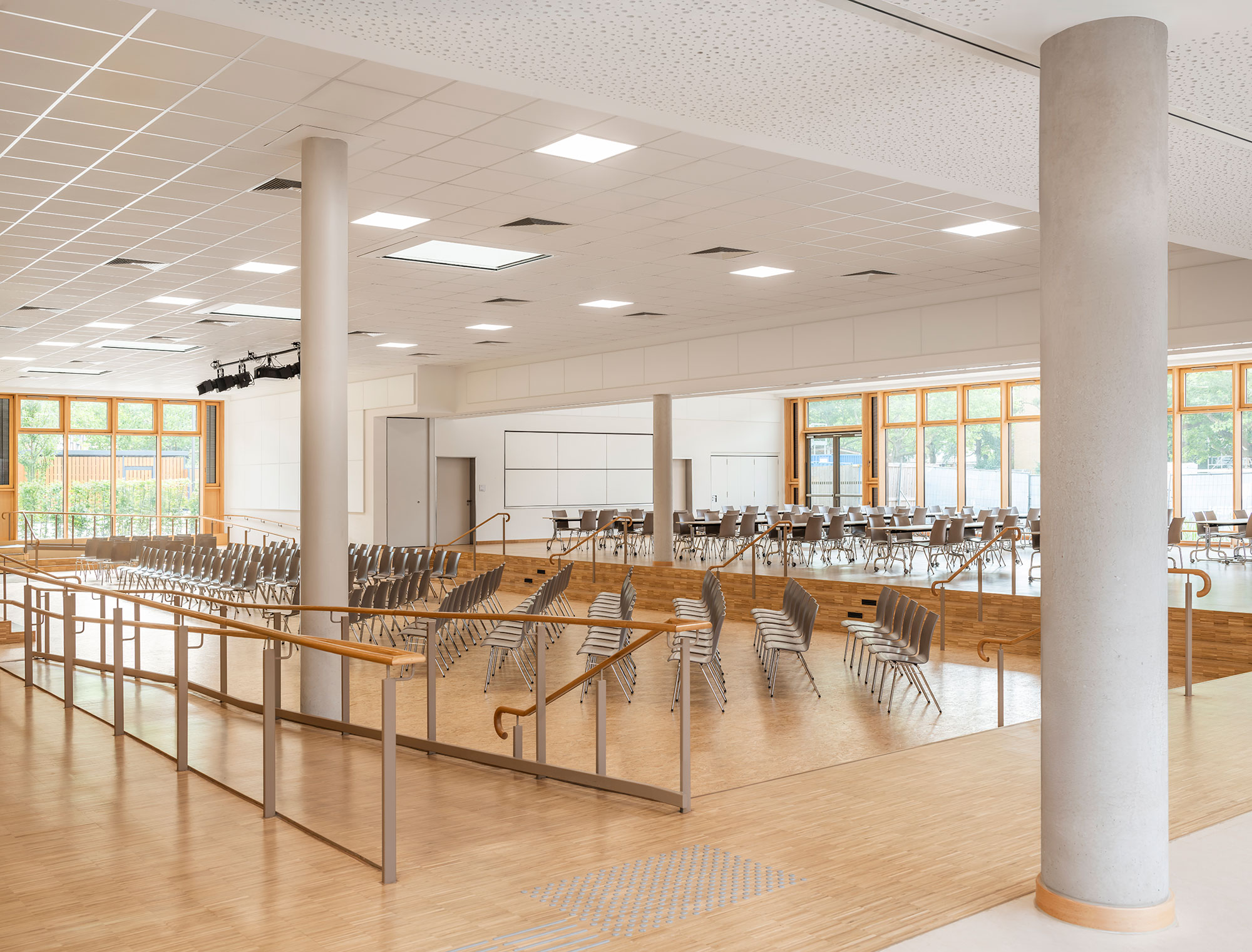 Großzügige Aula der neu errichteten Grundschule Mühlenberg mit heller Holzoptik, bodentiefen Fenstern und variabler Bestuhlung; im Raum befinden sich mehrere Ebenen mit Rampen und Geländern, eine offene Bühnenzone mit Beleuchtung sowie ein angrenzender Bereich mit Tischen und Stühlen für multifunktionale Nutzung.