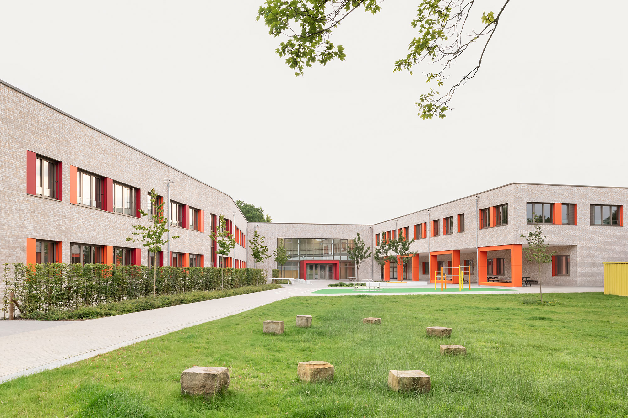 Weitwinkelansicht der neu errichteten Grundschule Mühlenberg, aufgenommen vom begrünten Außenbereich mit vereinzelten Findlingen als Sitz- oder Spielelemente; die zweigeschossige Klinkerfassade mit roten und orangenen Fensterakzenten umschließt den Schulhof U-förmig, während mittig der verglaste Eingangsbereich sichtbar ist.