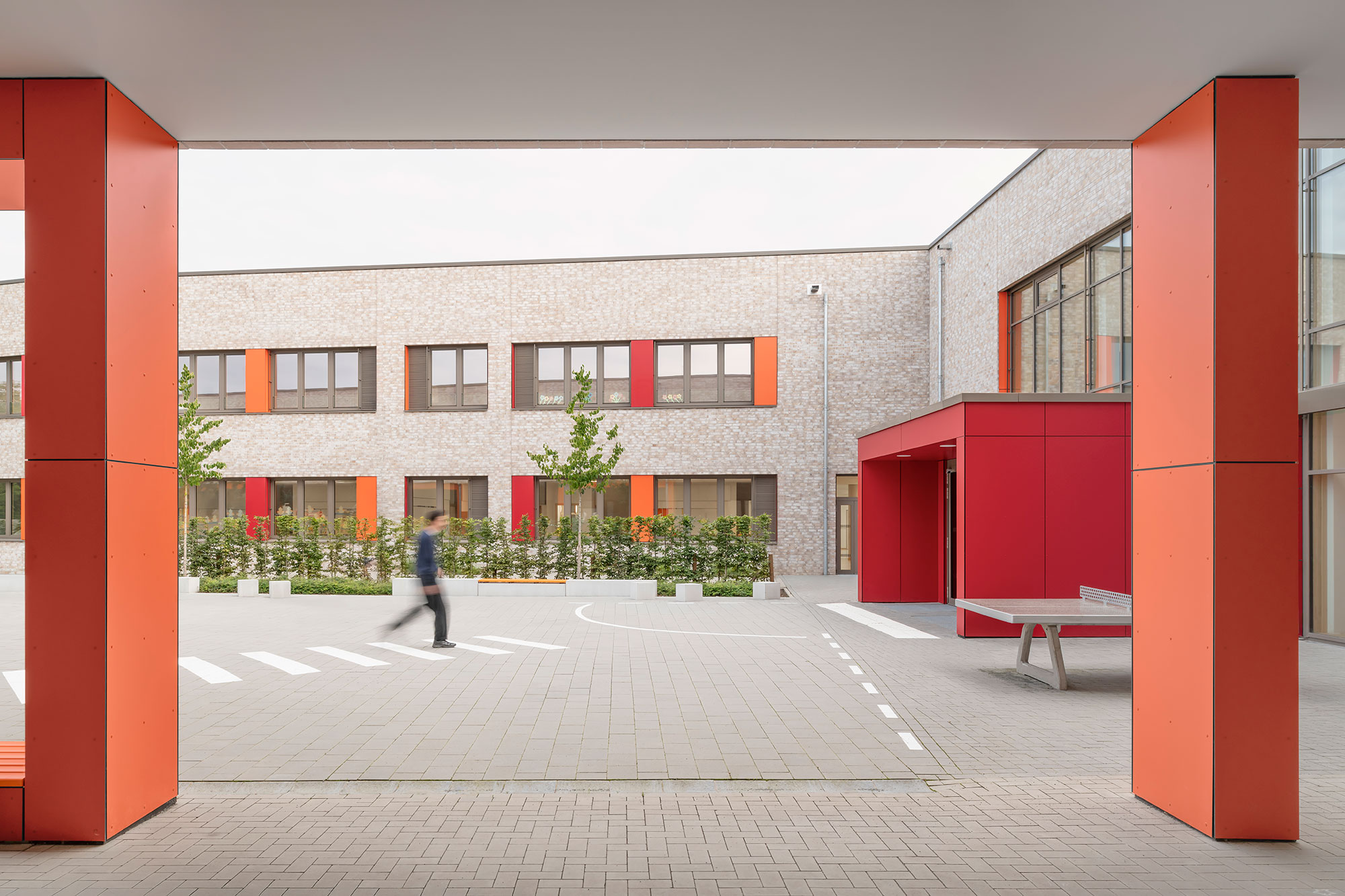Blick vom überdachten Bereich des Schulhofs auf den Innenhof der neu errichteten Grundschule Mühlenberg, mit rot-orangenen Fassadenelementen und Stützen, einer hellen Klinkerfassade, modernen Fensterreihen sowie einem bepflanzten Grünstreifen; im Vordergrund ist eine Person unscharf gehend zu sehen.