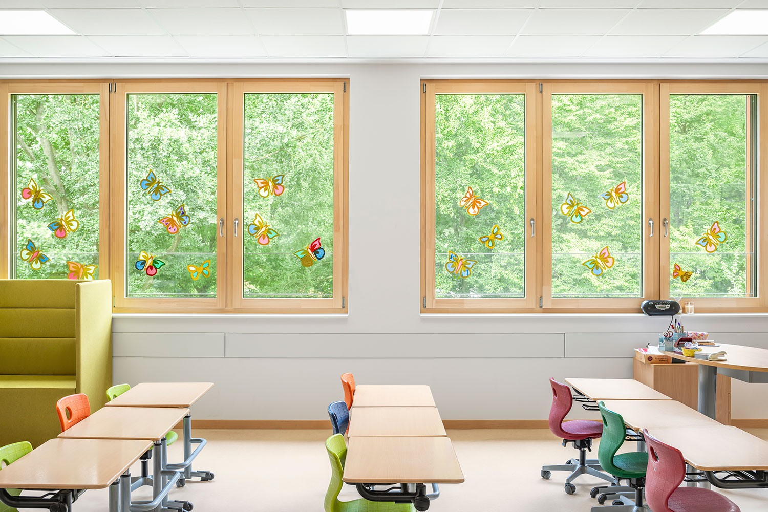 Heller Klassenraum der neu errichteten Grundschule Mühlenberg mit großen Holzrahmenfenstern, die auf dichten grünen Baumbestand blicken; die Scheiben sind mit bunten Schmetterlingsmotiven beklebt, davor stehen Tische und farbige Stühle der Schüler:innen sowie ein grünes Sitzmöbelstück für Rückzug und ein Lehrertisch mit Materialien.
