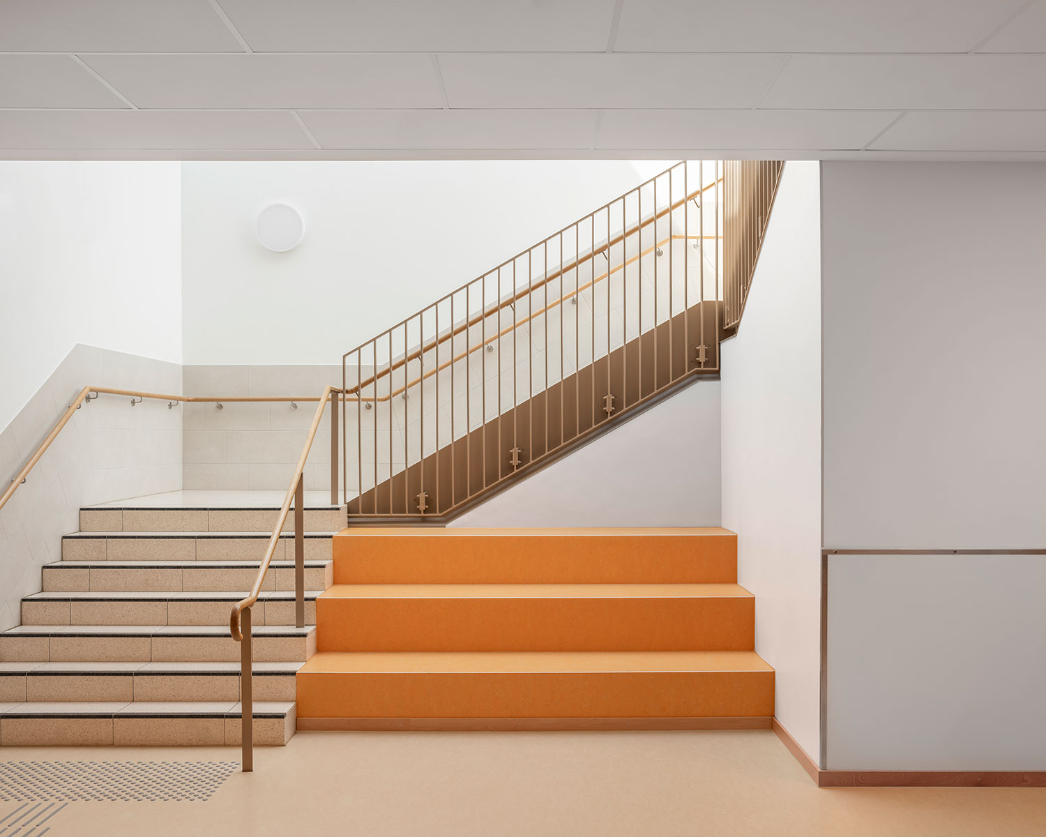 Treppenaufgang der neu errichteten Grundschule Mühlenberg mit hellen Fliesenstufen, orangefarbenen Sitzstufen im Vordergrund und einem warmtonigen Metallgeländer mit Holzlauf; die weißen Wände und das einfallende Oberlicht sorgen für einen freundlichen, lichtdurchfluteten Bereich.